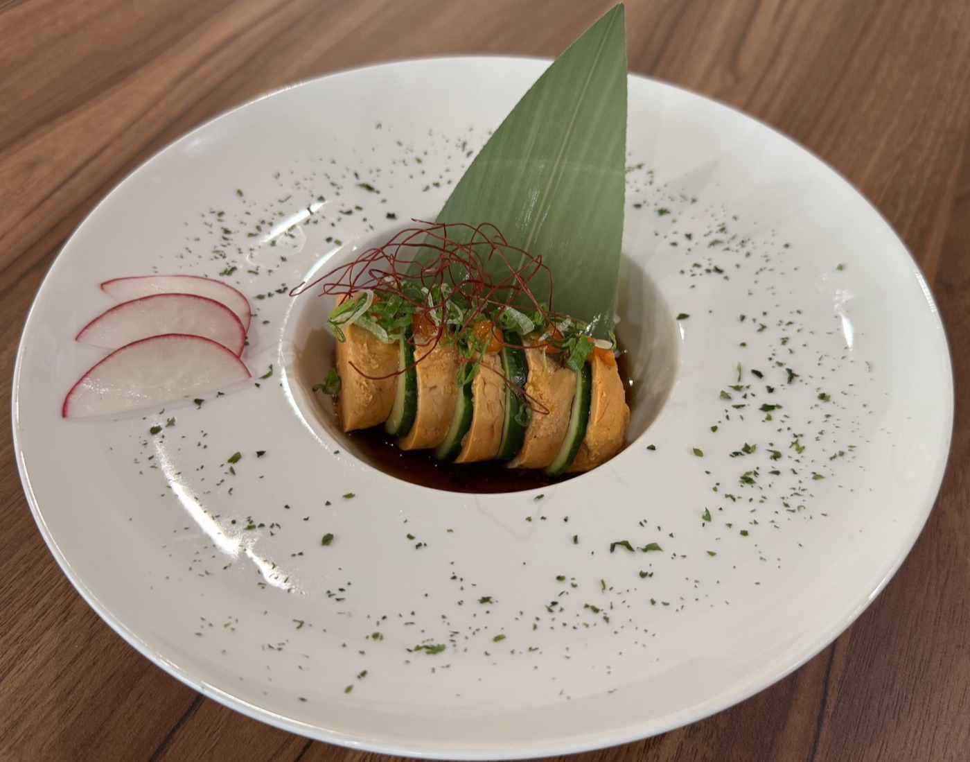 Orange and green slices in dark sauce, served in a sculptural white bowl with radish and a tall leaf