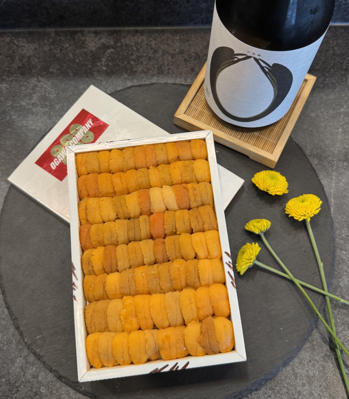 An open tray of sea urchin beside a sake bottle and yellow flowers on dark slate
